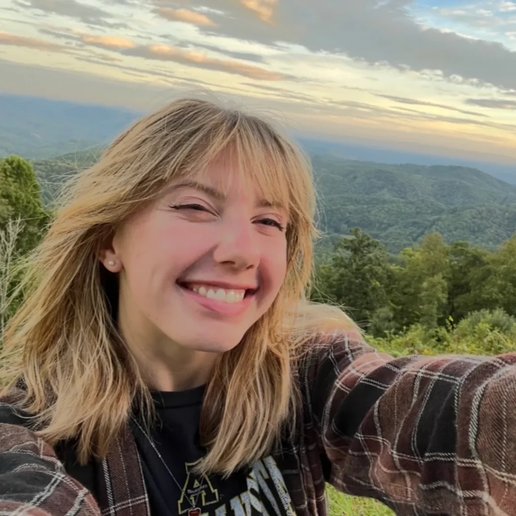 Smiling woman with blonde hair taking a selfie outdoors with scenic mountains in background.