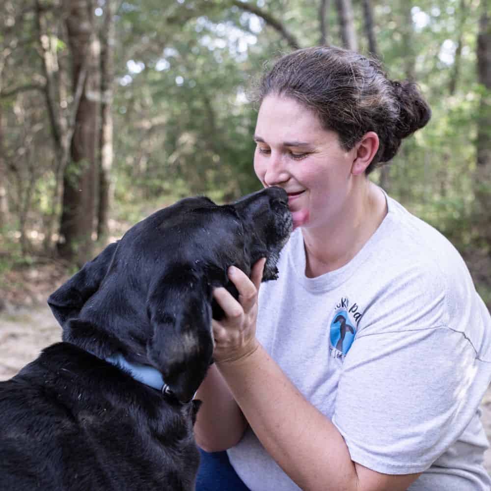 Woman and black dog sharing a loving moment outdoors in a wooded area.